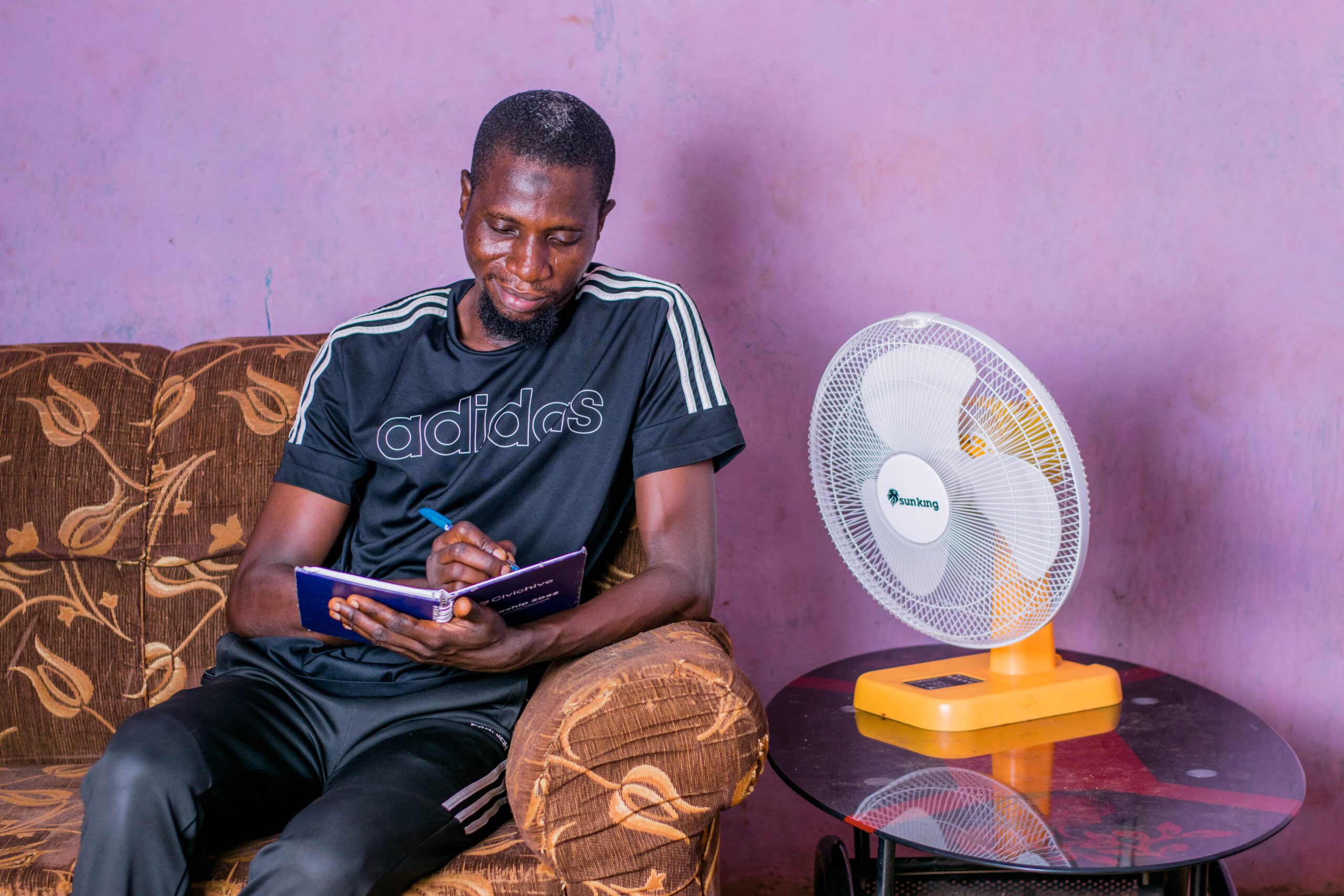 A man sits on a sofa reading. Next to him is the Sun King 16" Solar Table Fan, sitting on a small round table.